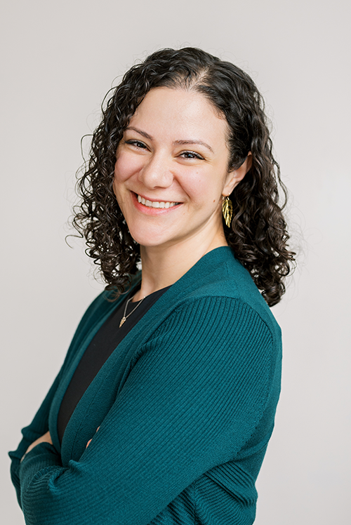 Headshot of Dr. Angela Izmirian Ph.D. smiling in a teal cardigan with her arms crossed in front of a grey wall.